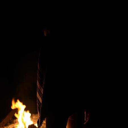 A Pinay woman is partially draped in a blanket, her figure outlined by the soft glow of the campfire. The blanket falls away on one side, revealing the curve of her hip and the silhouette of her body beneath. The darkness of the night contrasts with the light, hinting at what lies just out of view.