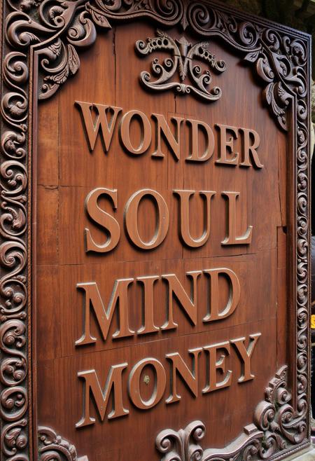 A large, ancient wooden sign, masterfully carved with exquisite and elaborate designs, evoking the feeling of a long-lost relic. The wood features deep, intricate patterns along the edges, including swirling vines, celestial symbols, and delicate filigree that resemble the craftsmanship of a forgotten civilization. Embedded in the wood are faint, mystical runes and nature-inspired carvings like leaves, flowers, and creatures, blending seamlessly with the deep grain and rich texture. The surface has a weathered look, with small cracks, knots, and areas where moss and lichen have naturally grown over time. Each letter is carved with precision in a hand-written, elegant font, flowing over four lines that read: ‘WONDER,’ ‘SOUL,’ ‘MIND,’ and ‘MONEY.’ The letters are adorned with tiny decorative accents, as though they are part of the overall design, merging with the wood’s ancient, sublime artistry. The whole piece radiates a sense of mystery and timeless beauty, as if it holds ancient wisdom within its carved surface