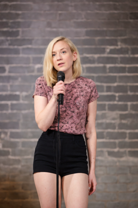 1girl, Female stand-up comedian, holding a microphone, performing on stage, casual attire, confident expression, spotlight, brick wall background
 <lora:emily_kinney_lora_v02:1> ek84, blonde hair