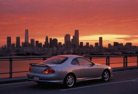 (1996 Hyundai Tiburon RD) motor vehicle, no humans, city, sky, outdoors, scenery, orange sky, road, building, cityscape, sunset, vehicle focus, skyscraper, sports car, shadow, gradient sky, red sky, evening, cloud, street, skyline, twilight, spoiler \(automobile\), orange background, dusk, red background, water, solo, bridge,