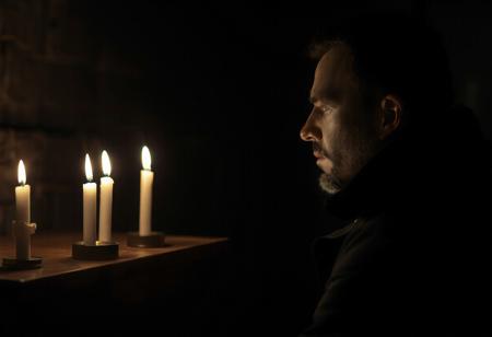 professional photo, side view photo of Caucasian man in kneeling in front of an altar with 3 lit candles on it, watching the 3 candles, wearing a dark blue parka, serious face, contemplative expression, dark features, dramatic shadows, very dark and underexposed, face lit only by candles, all else dark