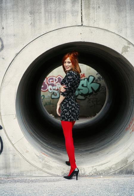 Photograph of a beautiful 20 year old redhead woman with shoulder-length hair in a loose bun. She is wearing a short, tight black print dress, red leggings, and black patent leather shoes with low heels. She is standing inside a large concrete storm culvert set into a concrete wall. The wall is covered with graffiti. Viewed from the side, her head turned toward the viewer. She is smiling at the viewer with her mouth closed. Film photo, film grain, professional photo shoot.