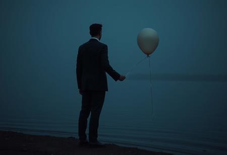 Man in a three piece business suit standing at the shore of a lake facing away from the viewer. He holds a balloon by a string in his right hand. The balloon isn't floating but has sunk to the ground. dark image, dim light, sunk balloon, three piece business suit, man standing, lake, realistic photo, raw photo, hdr, 8k, highly detailed
