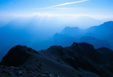 Immerse in a breathtakingly beautiful landscape: a vibrant blue-hued horizon stretches across the frame, fading into an atmospheric haze. Unusual dark colors, such as deep indigo and rich charcoal, punctuate the scene with dramatic flair. Cinematic lighting casts long shadows, emphasizing the fine details of the terrain. Film grain adds texture to the image, while superb composition guides the viewer's eye through the masterpiece. Maximum detail is achieved in every element, from the rugged terrain to the wispy clouds, creating an unparalleled level of visual mastery.