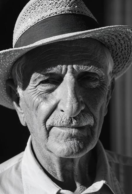 B&W headshot, a 75yo italian man, light filtering through a hat's brim, casting intricate shadows on the face