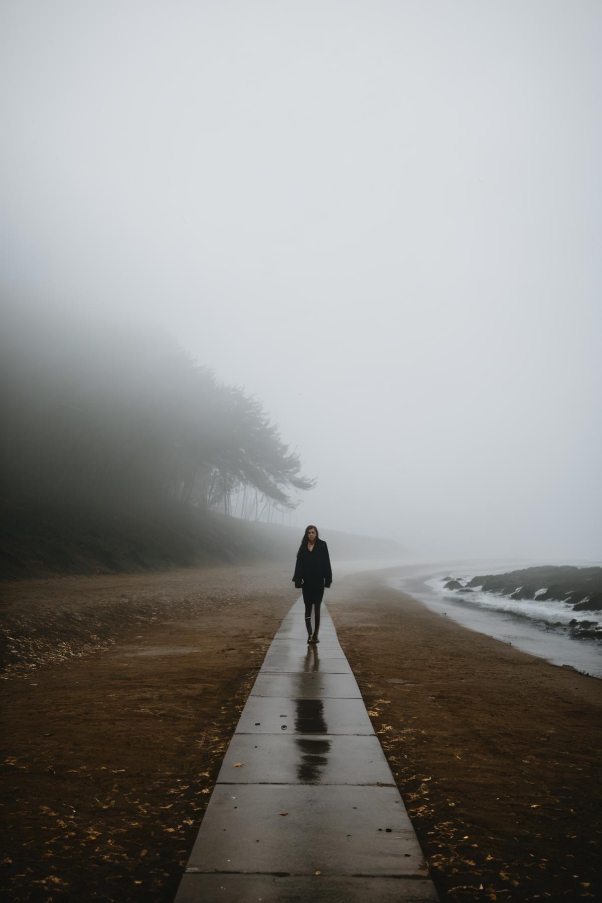 A woman walking on a path near the water in a foggy environment.