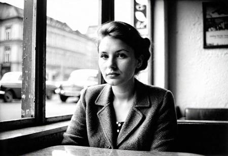 german woman 30 years old sitting in a cafe in east berlin in 1962, watching outside, face in shadow, low light, full body shot from side, 1960 soviet look, dark mood, ilford hp5 plus 400, medium format camera,artsy, shadows, wide angle, contact test, damaged print, faded, grainy,blurry, pixelated, motion blur, vignette, high contrast