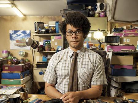 Richard Ayoade as Moss, parted afro, plaid shirt, paisley tie, portrait stock photo middle-aged man in cluttered office, basement office, sitting at desk, (masterpiece:1.2) (photorealistic:1.2) (bokeh) (best quality) (detailed skin) (intricate) (8k) (HDR) (cinematic lighting) (sharp focus) (Clutter-Home:1.2) <lora:Richard-Ayoade-v10:0.7>