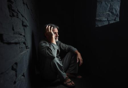 a dark room with walls and floor of solid black stone. the room is damp and cold and dark. a man sits against a wall on the left side staring ahead toward the right side of the photo. his hand is raised to his head holding his head and running his fingers through his hair. he wears old and tattered clothes. he has dark circles under his eyes to show his weariness. his hair is messy. he looks aged from the struggle of his life. it is a dark and grim image that portrays the depth of pain felt by depression.
