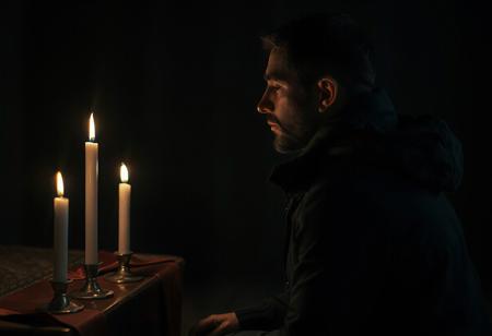 professional photo, side view photo of Caucasian man in kneeling in front of an altar with 3 lit candles on it, watching the 3 candles, wearing a dark blue parka, serious face, contemplative expression, dark features, dramatic shadows, very dark and underexposed, face lit only by candles, all else dark