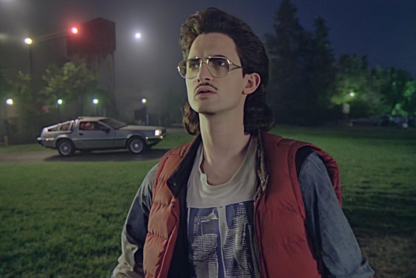 A man wearing glasses and a red vest standing next to a DeLorean.