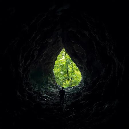 Dark, jagged cave interior with protagonist slowly advancing toward a small opening revealing lush green foliage outside, harsh contrast between shadowy confinement and bright natural world representing hope and escape.