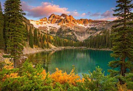 A breathtaking autumn landscape featuring a tranquil mountain lake reflecting a rich tapestry of foliage. The background consists of majestic mountains with snowy peaks, basking in the glow of a sunset that casts a warm orange light on the cliffs. The mountainsides are densely forested with various shades of green conifers, transitioning into a vibrant array of autumnal colors such as yellows, oranges, and reds as the elevation lowers. The near bank of the lake features lush vegetation with a multitude of colorful plants and bushes, interspersed with scattered rocks. A serene and crystal-clear lake at the center mirrors the beauty of the surrounding nature, doubling the visual impact of the foliage and the majesty of the mountains. The sky above is a gradient of soft blues and greens, suggesting either dawn or dusk. The composition should elicit a sense of peace and the crisp freshness of mountain air.