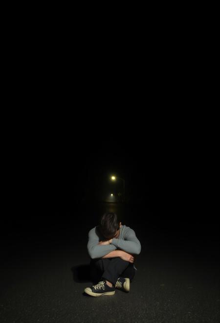 it is night. there is no light aside from the distant street light. the street is empty. the homes along the street are all dark. tall imposing trees stretch over the dark street. a man sits with his knees pulled up to his face and his arms wrapped around his legs. his face isn't visible but it seems he is sad. he sits in the street. I can not express enough how dark it is in this image and how upset I will be if it isn't dark.