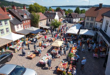 small town Saturday market day, sunny, northerly, July, crisp, near the sea,, village square filled with 40 tables, 20 booths, crafts, homemade food, carvings, buyers, sellers, colorful, chocolate sop, bakery, parked cars, produce, berries, pottery, wool weaving, hot dog grilling, bookstore, bank building, tea room, view from above