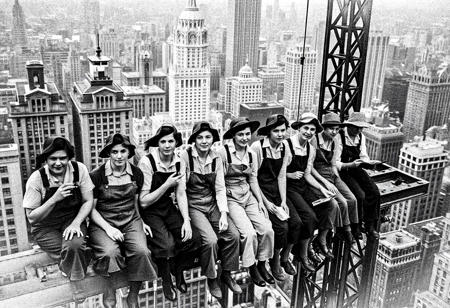 This black-and-white photograph captures a group of ten women sitting on the steel beam of a skyscraper, likely during the construction phase of the building. The women are dressed in work clothes typical of the early 20th century, including overalls, shirts, and caps, with some wearing suspenders. They appear to be taking a break, with most holding cigarettes and some holding newspapers or books. Their expressions range from relaxed to contemplative. The women are seated in a row, most are not facing the camera, and are evenly spaced along the beam. The background reveals a dense, urban cityscape with numerous tall buildings, indicating a metropolitan area. The buildings have a mix of architectural styles, with some featuring large, flat rooftops and others with more intricate designs. The photograph captures a moment of camaraderie and labor among the construction workers, highlighting the physical and mental strain of their work. The image's historical context is evident, as the photograph likely dates to the early 1900s, when skyscrapers were being built at a rapid pace. The black-and-white format adds a timeless quality to the photograph, emphasizing the stark contrasts and textures of the scene.