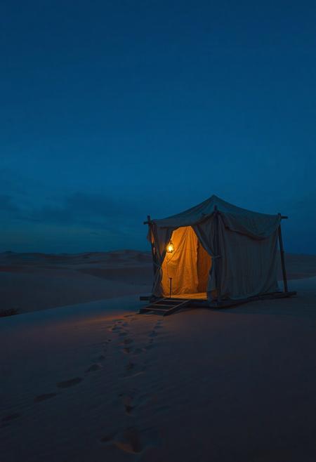A nostalgic 4K desert scene at golden hour — endless dunes bathed in deep blue twilight, soft mist hovering near the horizon. A weathered caravan stands still, its fabric gently fluttering in the breeze. Faint lantern light flickers inside, casting long shadows on the sand. Footprints trail off into the distance. The air is still, heavy with memories. Perfect for a slow dolly shot — cinematic framing, emotional atmosphere, deep focus, soft grain, One Heart style, ultra-detailed, warm and cool tones blending in harmony.