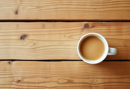 A view from the top of a wooden table with a coffee mug, classic stock photo, chill vibe, latte cafe on a classic wooden kitchen table. Empty table except the coffee mug. Coffee mug on the far right. Clean mug with fresh coffee, light foam on coffee. No space between plank, glued plank without gaps. Light coloured wood
