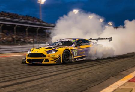 "This image portrays a race car in full motion on a track, with a cloud of smoke trailing behind it. The car's striking yellow and purple color scheme, along with black accents, creates a visually captivating appearance. The smoke adds a sense of speed and intensity to the scene. The well-lit track and the enthusiastic crowd in the stands contribute to the excitement and intensity of the race."