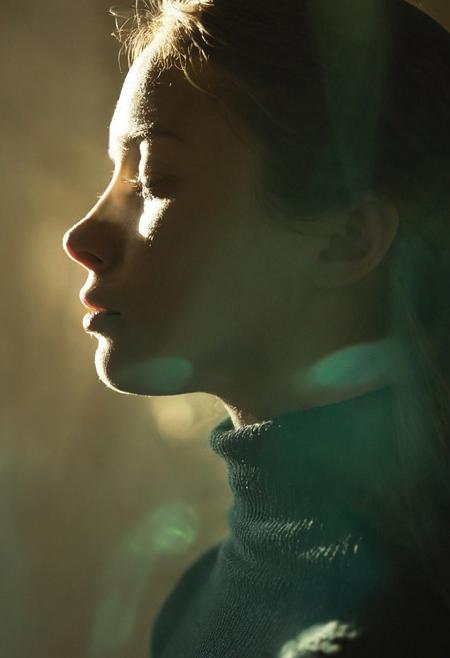 A dreamy portrait photograph captures a young woman in perfect right-facing profile, illuminated by natural dappled sunlight that creates intricate patterns of light and shadow across her face and neck. The image employs shallow depth of field and warm tones, with the subject wearing a dark high-necked garment against a soft, out-of-focus background in muted teal and beige tones. The composition emphasizes the interplay between light and shadow, creating an ethereal, atmospheric aesthetic enhanced by the golden hour lighting conditions. Portrait photography with shallow depth of field and natural lighting.