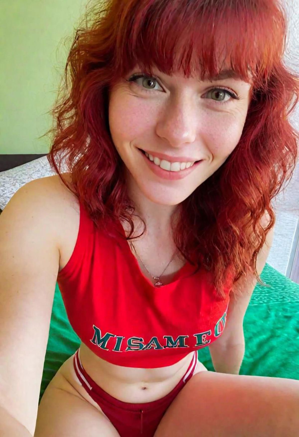 A young woman wearing a red shirt and underwear, smiling for the camera.