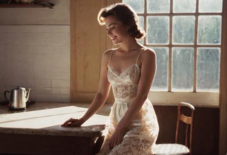 grainy 1950s amateur smut color photograph, full body profile photograph of a cute housewife wearing a satin slip with spaghetti straps and lace trim, leaning over a kitchen table, (morning sunlight creates a silhouette of her breasts through her silky satin slip), outline of boobs and nipples in silky satin slip, backlight, small boobs, wide hips, upper thigh, very detailed, angle from side, silhouette, sun rays, profile, detailed eyes and face, cute smile, film grain
