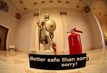 “Create an image of a statue of Giovanna d’Arco (Joan of Arc) displayed in a museum setting, shot using a Hasselblad camera with 12mm fisheye optics. The statue should have a classic, realistic style, capturing Joan in her iconic armor, holding a sword and shield, but with a gentle, subtle smile on her face, softening the heroic pose. The setting is an elegant, spacious museum with marble floors and soft lighting highlighting the statue. Near the base of the statue, there should be an extinguisher (extintor) subtly placed in the scene, offering a contrast between the historical figure and the modern, functional object. In the foreground, include a small panel with the text ‘Better safe than sorry!’ in bold, plastic extruded characters, with a 3D glossy plastic finish. The fisheye lens should distort the perspective slightly, creating a dynamic, wide-angle view that enhances the contrast between the ancient statue and the modern safety feature, adding a surreal and playful touch to the composition.”