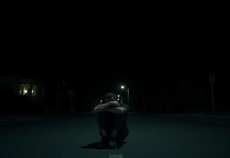 it is night. there is no light aside from the distant street light. the street is empty. the homes along the street are all dark. tall imposing trees stretch over the dark street. a man sits with his knees pulled up to his face and his arms wrapped around his legs. his face isn't visible but it seems he is sad. he sits in the street. I can not express enough how dark it is in this image and how upset I will be if it isn't dark.