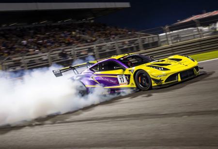 "This image portrays a race car in full motion on a track, with a cloud of smoke trailing behind it. The car's striking yellow and purple color scheme, along with black accents, creates a visually captivating appearance. The smoke adds a sense of speed and intensity to the scene. The well-lit track and the enthusiastic crowd in the stands contribute to the excitement and intensity of the race."
