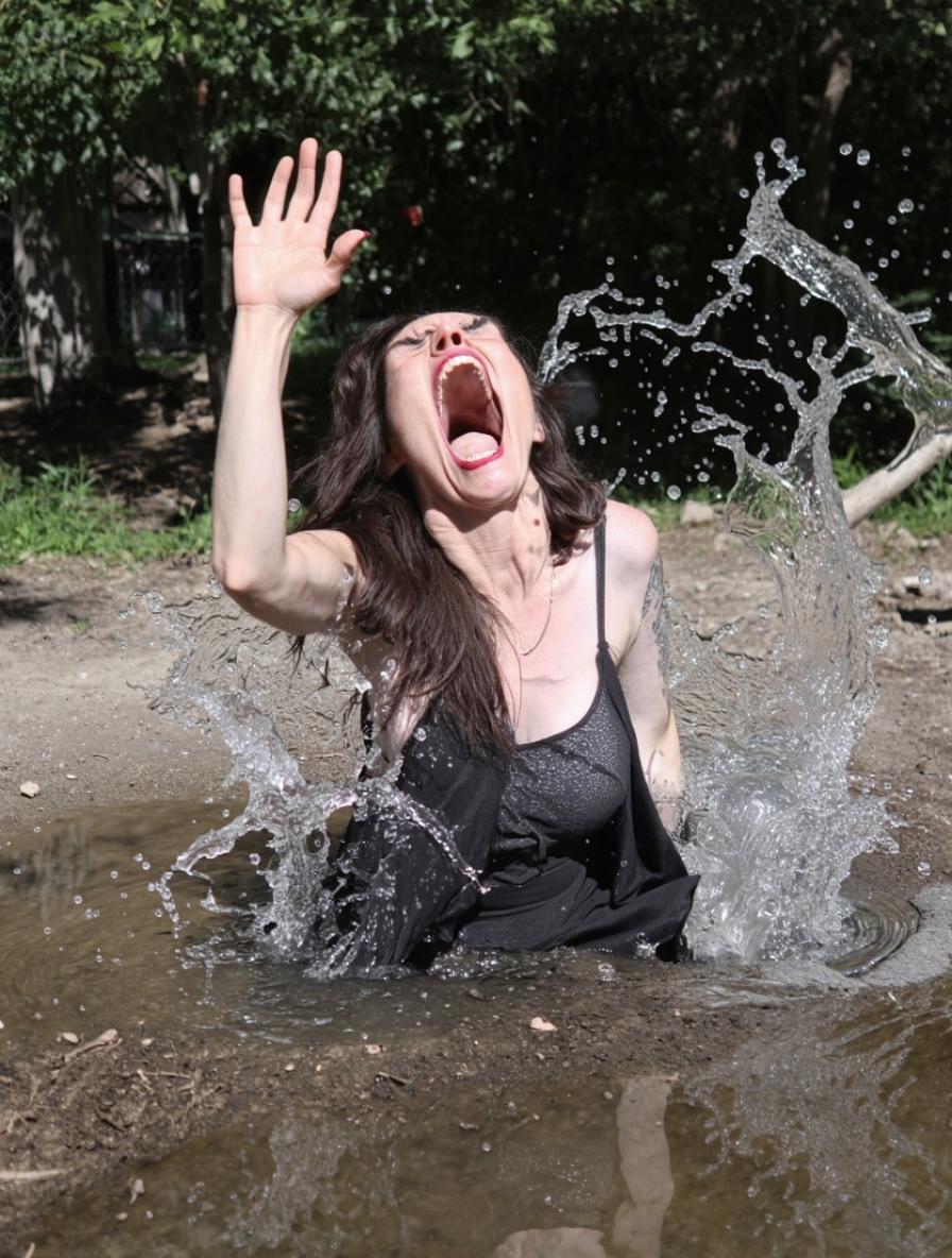 iph0ne amateur photo of a hysterical woman, screaming and tantrum and drowming on a small puddle of water, splash splatter, closed eyes, park on the background, raised hand asking for help, 35mm photography