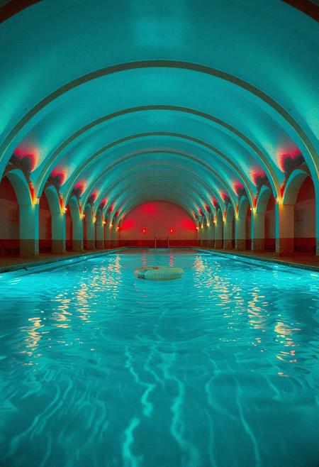 "An endless, looping pool complex with glowing teal-blue water, perfectly still yet echoing with distant, impossible splashes. The vaulted archways repeat into infinity, their neon-lit edges reflecting off the wet concrete in oily rainbow smears. The air is thick with chlorine-scented mist, distorting the flickering overhead lights. The water is crystal clear but too deep—something moves just beyond visibility, stirring the ripples unnaturally. No exits, no people—only a single abandoned pool float drifting towards the viewer. Shot on grainy 35mm film with a fisheye lens, the edges of the frame warped like a VHS tape left to decay. Vaporwave aesthetics meet psychological horror—the colors are vibrant but sickly, the geometry almost correct. Submerged shadows suggest elongated limbs just below the surface.
