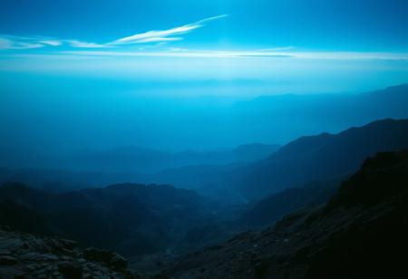 Immerse in a breathtakingly beautiful landscape: a vibrant blue-hued horizon stretches across the frame, fading into an atmospheric haze. Unusual dark colors, such as deep indigo and rich charcoal, punctuate the scene with dramatic flair. Cinematic lighting casts long shadows, emphasizing the fine details of the terrain. Film grain adds texture to the image, while superb composition guides the viewer's eye through the masterpiece. Maximum detail is achieved in every element, from the rugged terrain to the wispy clouds, creating an unparalleled level of visual mastery.