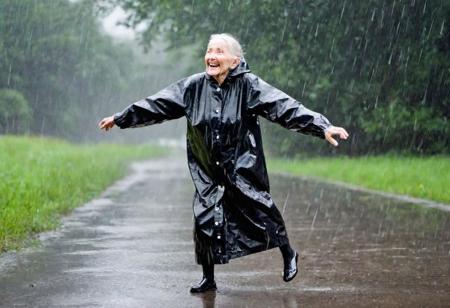 An 80 year old elderly woman wears a very tight fitting puffy black ankle length raincoat made of PVC, she is very happy as rain droplets appear on her jacket in a light rain shower