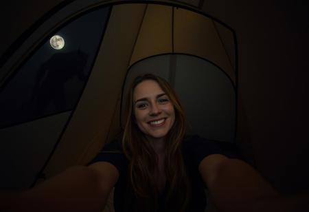 A woman taking a selfie inside a small modern tent at night. She is smiling for the camera and winking. Behind her is the wall of the dark tent illuminated by the camera flash. In the top left of the image the sheer and partially see-through mesh of the tent is visible. A scary disturbing eerie creature of some sort is barely visible through the mesh in the top left of the image and is leaning over the outside of tent. The woman taking the selfie is oblivious to its presence since it is outside and not easily visible in the image. The creature is tall and looks like a wendigo cryptid and very dimly lit by moonlight but is primarily visible via its silhouette. The woman is in the center of the image and is the focus, not the creature.