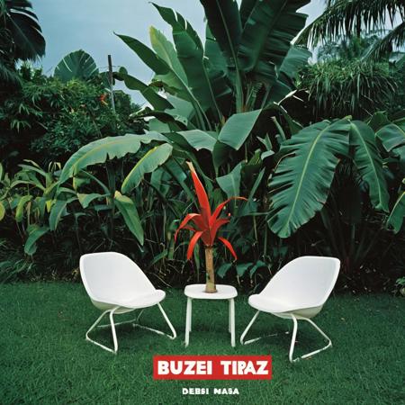 This is a photograph of the cover of an album "DeBÍ TiRAR MáS BUZZ". On the left, two white plastic chairs are placed on a patch of grass, partially obscured by lush greenery. The background includes large banana leaves and tall plants. The album cover on the right is predominantly blue with bold red and white text. It features a lightning logo and various tracks listed, including "BUZZ" and "BUZZ BUZZ." The overall aesthetic is vibrant and tropical.