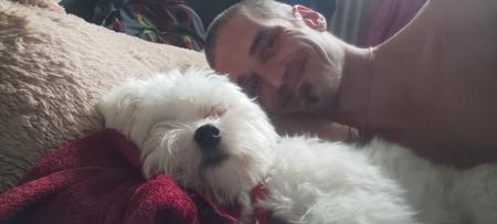 Godmaster laying in bed smiling, cuddled up to a little white dog 