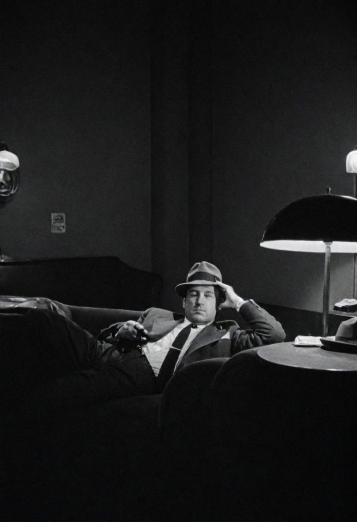 A man with a hat and tie is sitting on a couch in a dark room.