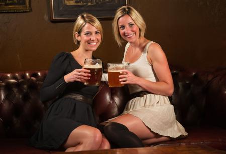 (2girls) two 30-something women, close friends, linen dresses, (one blonde and one brunette, short hair) each with a pint glass of  English pale ale (IPA, real ale, traditional ale),  sat in an old-style English pub, Victorian leather-bound furnishings, natural light, ambient lighting.