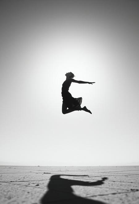 A figure mid-leap against a bright, empty sky, where their body is frozen in time, resembling one of’ motion-filled yet impossibly elegant compositions, but subtly altered by a surreal touch.surreal minimalist photography with high-contrast sculptural elegance, black-and-white dreamlike atmosphere, dramatic light and shadow interaction, faceless figures blending into vast empty spaces, poetic movement frozen in time, timeless yet surreal storytelling