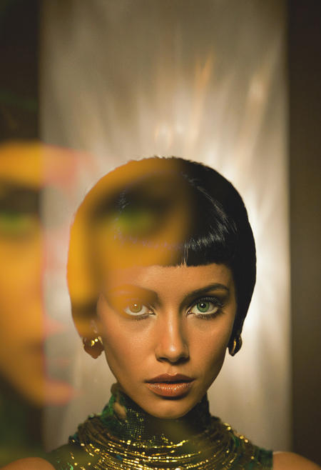 A stunning portrait of a woman embodying Cleopatra—short, sleek black hair, piercing emerald-green eyes, and flawless golden-brown skin illuminated by a flickering candle’s glow. The golden light dances across her sharp cheekbones and full lips, casting ephemeral shadows that shift like whispers. She wears a contrast-rich garment (deep crimson or black with gold accents), the fabric’s texture barely visible in the shallow depth of field. The background dissolves into velvety darkness, emphasizing her hypnotic gaze. Shot with a painterly chiaroscuro style, rich amber tones melting into inky blacks, and a textured film grain overlay for a cinematic, timeless feel. 35mm film aesthetic, subtle noise, hyper-detailed skin texture, and an air of regal mystery