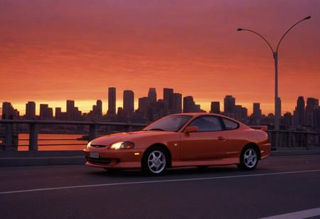 (1996 Hyundai Tiburon RD) motor vehicle, no humans, city, sky, outdoors, scenery, orange sky, road, building, cityscape, sunset, vehicle focus, skyscraper, sports car, shadow, gradient sky, red sky, evening, cloud, street, skyline, twilight, spoiler \(automobile\), orange background, dusk, red background, water, solo, bridge,