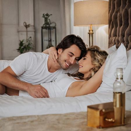 The photo captures the moment when the happy young couple is in a luxurious bedroom. A 25-year-old European man with dark brown shoulder-length hair falling on his shoulders and neck lay down in bed and tenderly and lovingly hugged his beloved woman. A 23-year-old woman with long wavy blonde hair is lying in bed . The composition focuses on the couple, 
emphasizing their tender relationship.