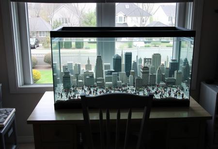 A large bustling city is in a glass aquarium on a desk with an empty chair pushed in under the desk.  The desk is in front of an open window.  A suburban yard with ornamental trees and a street with houses across the street can be seen out the window.
In the aquarium, tiny people can be seen going about their normal everyday lives.