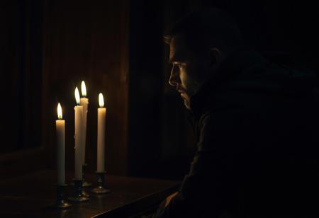 professional photo, side view photo of Caucasian man in kneeling in front of an altar with 3 lit candles on it, watching the 3 candles, wearing a dark blue parka, serious face, contemplative expression, dark features, dramatic shadows, very dark and underexposed, face lit only by candles, all else dark