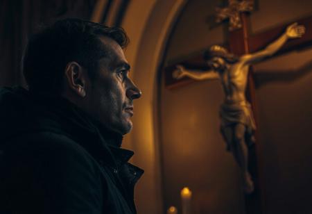 professional photo, side view photo of Caucasian man wearing a dark blue parka with dark features and a serious contemplative expression looking up at a plaster statue of Jesus on the cross mounted on the wall of a church, the man and Jesus are shown in profile, the scene is very dark and underexposed with only the man's and Jesus' face lit by warm, dim candle light, everything else is dark shadows